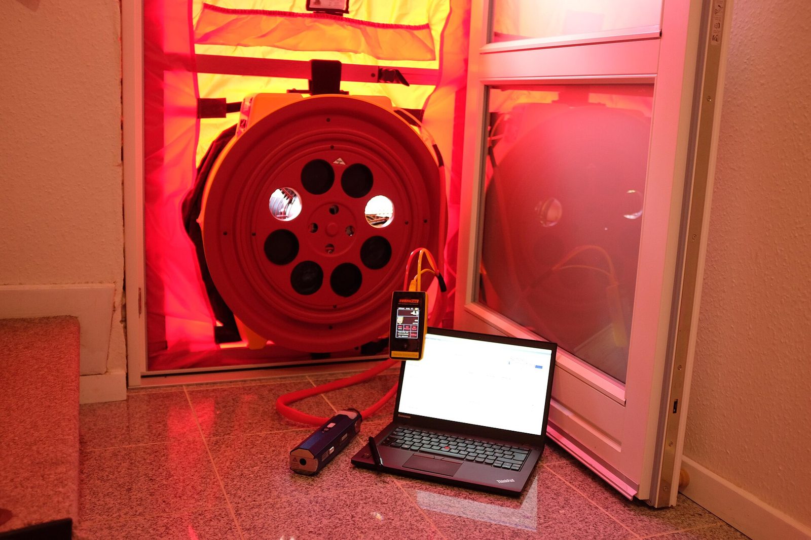 Luchtdichtheidstest met een rode ventilator in een deuropening, aangesloten op een laptop op de vloer
