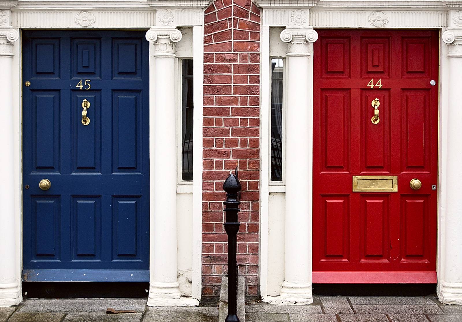 Twee voordeuren naast elkaar, een donkerblauwe en een rode, met koperen kloppers tussen witte zuilen